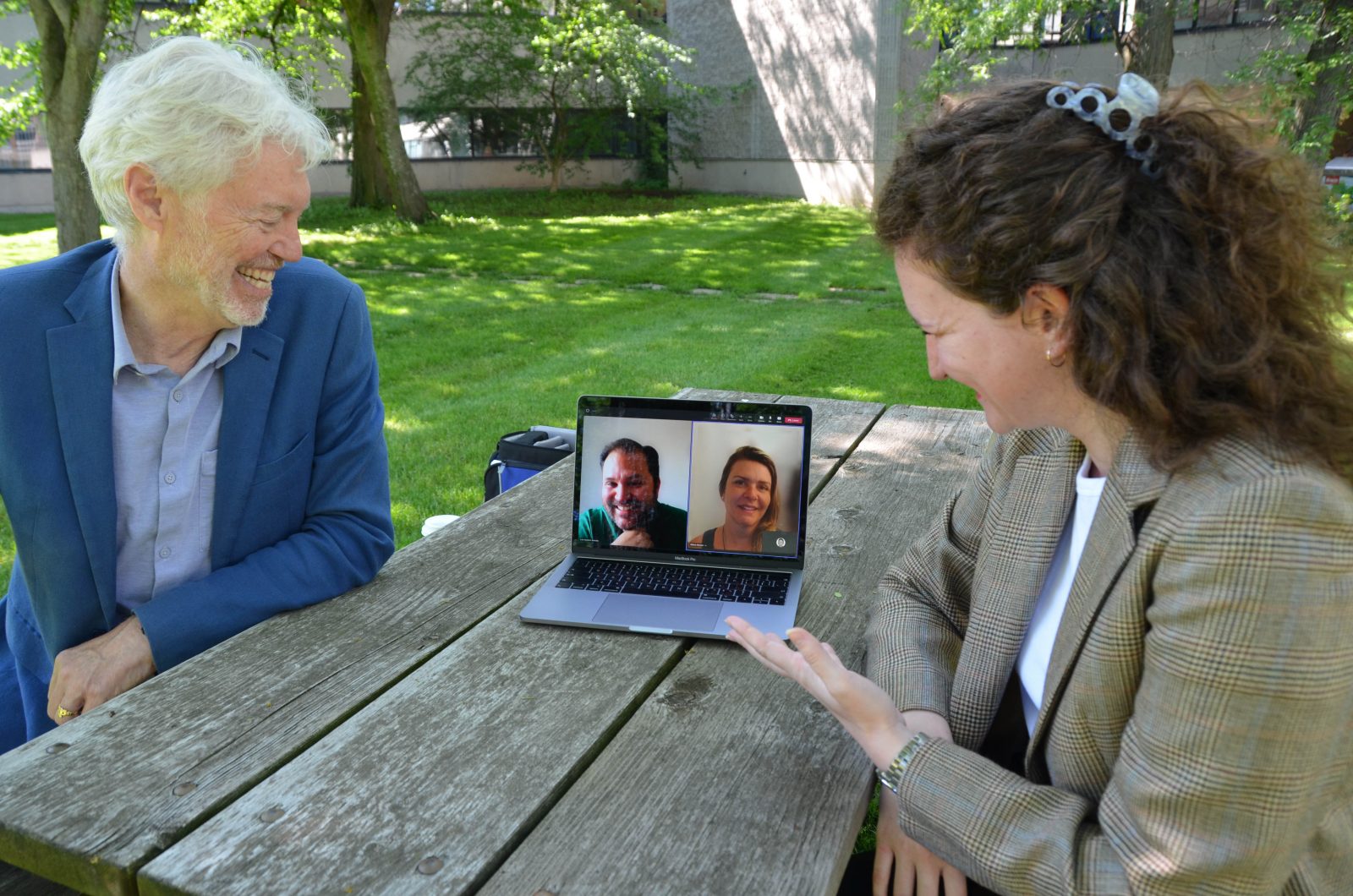 A laughing man with white hair wearing a medium blue jacket and a light blue shirt sitting at a picnic table leans in to look at a laptop screen with a man and a woman’s faces on the screen while a smiling woman with long brown hair, a brown checkered blazer and a white top and gesturing with her hand is also leaning into the laptop, with green space and a building in the background.