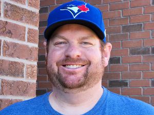 A portrait of David Ling, wearing a blue T-shirt and Toronto Blue Jays baseball cap and standing in front of a red brick wall.