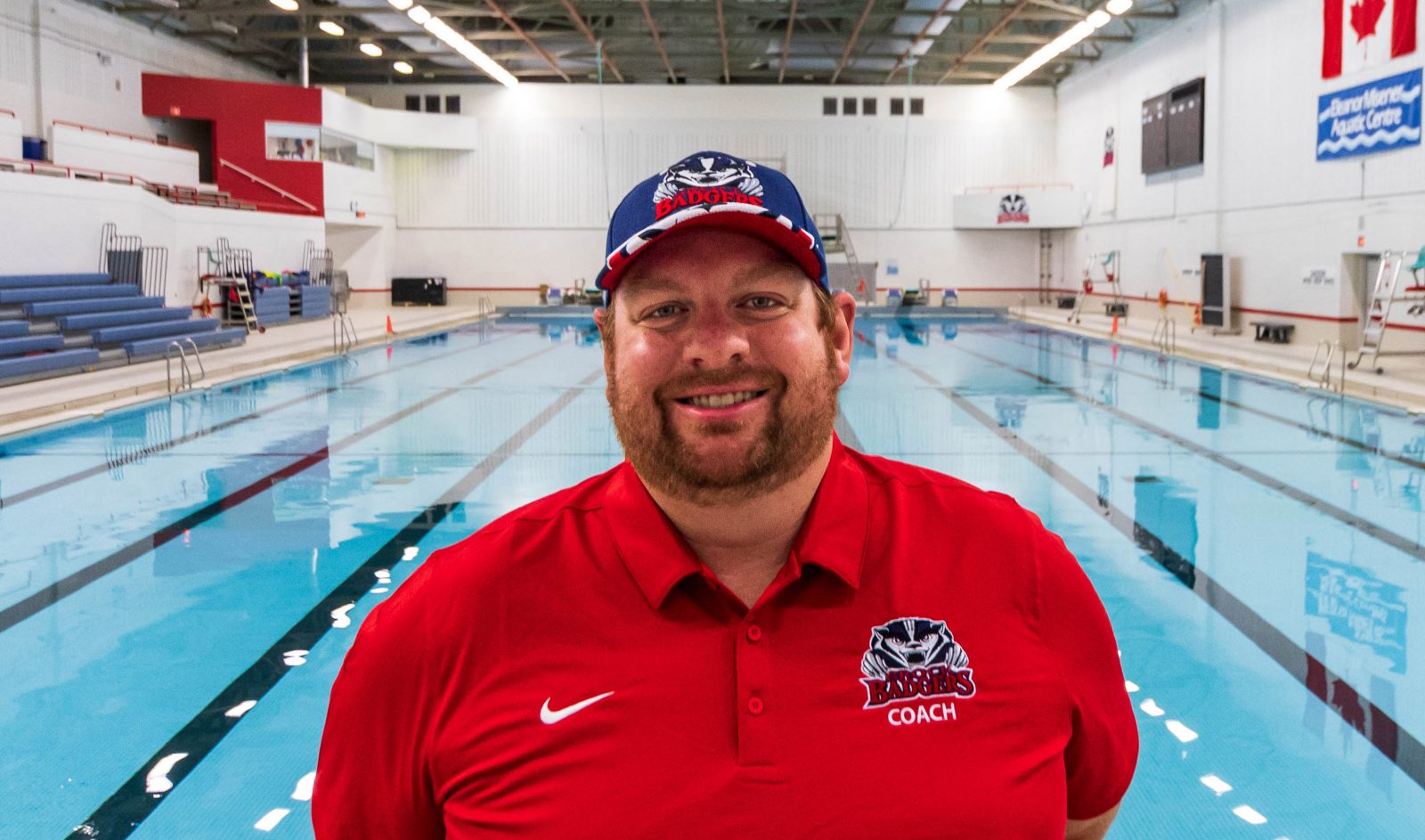 A man in a red Brock Badgers polo shirt and blue Brock Badgers baseball cap stands in front of a swimming pool.