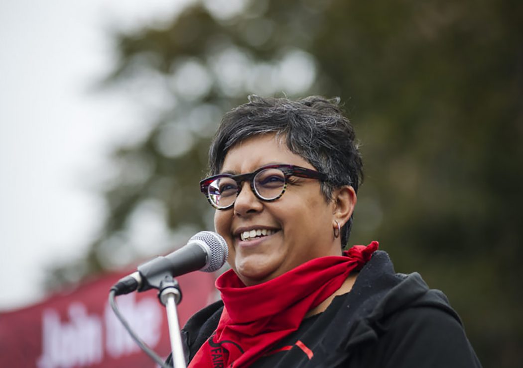 A woman with a red scarf and black glasses stands at a microphone.