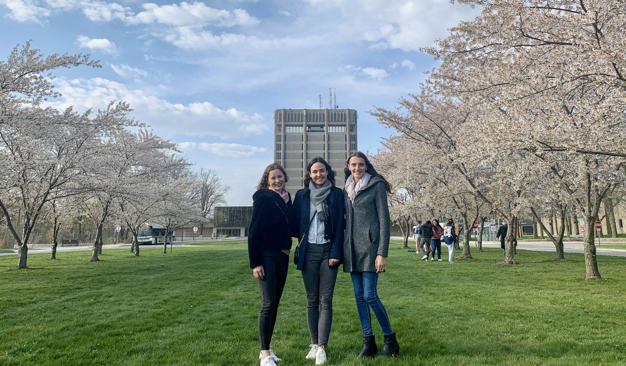 Three students stand in front of Brock’s Schmon Tower with cherry blossom trees in bloom on either side.