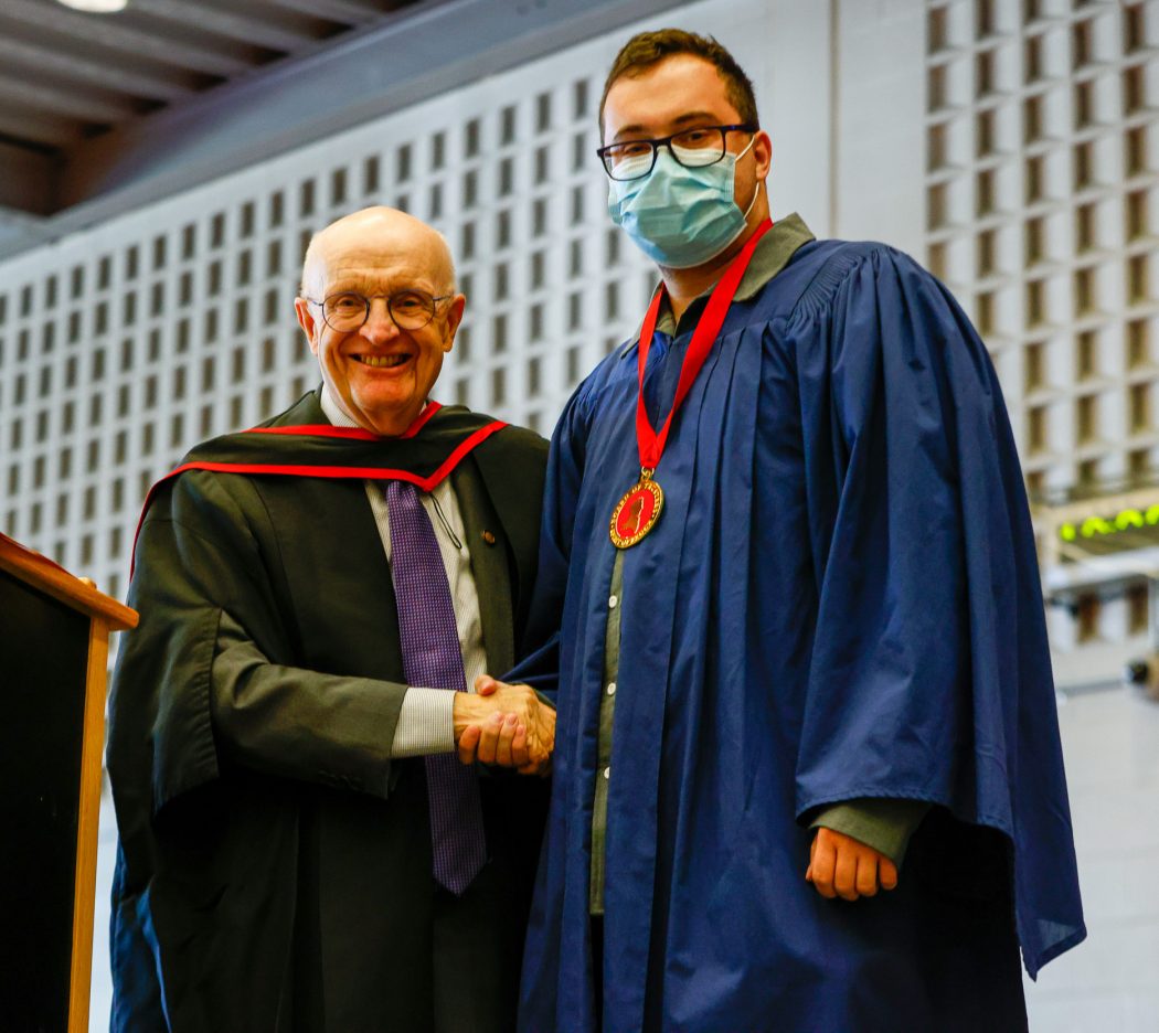 Two men in graduation gowns stand together on stage smiling.