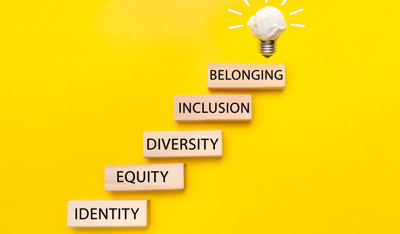 Building blocks that read 'identity,' 'equity,' 'diversity,' 'inclusion,' and 'belonging' form a staircase that ends in a lightbulb sculpture against a yellow background