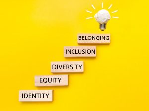 Building blocks that read 'identity,' 'equity,' 'diversity,' 'inclusion,' and 'belonging' form a staircase that ends in a lightbulb sculpture against a yellow background