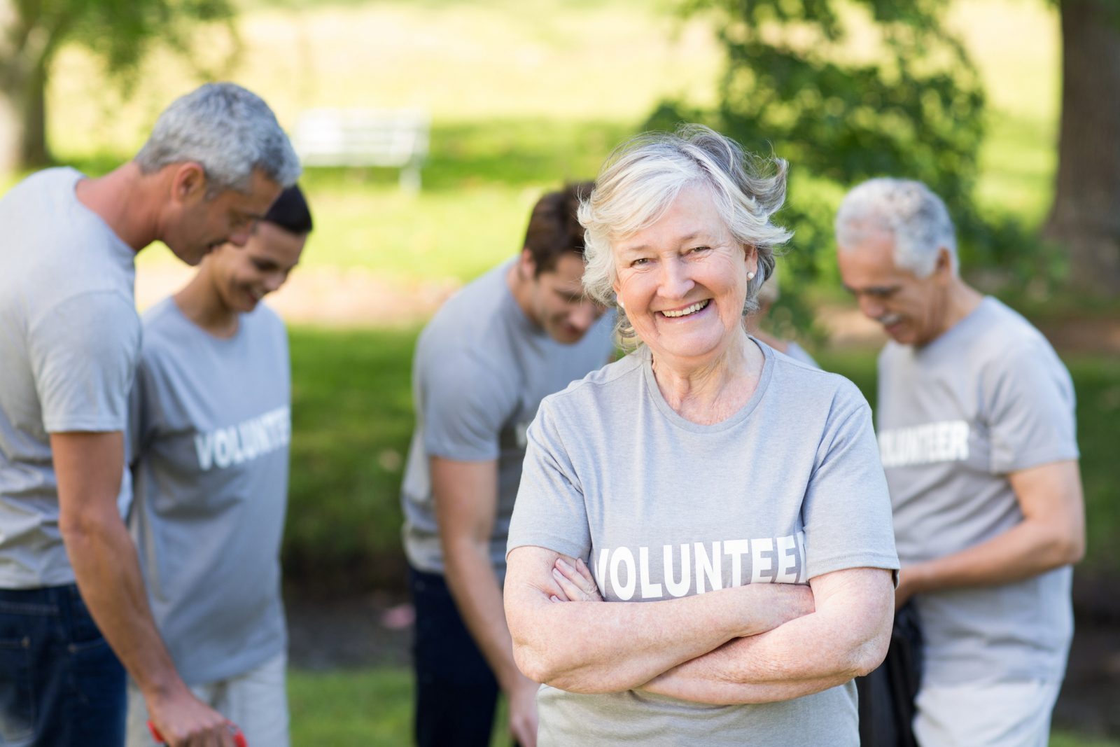 A senior woman in a grey shirt with the word 'VOLUNTEERS' written in white letters across the front, stands in front of four other volunteers.