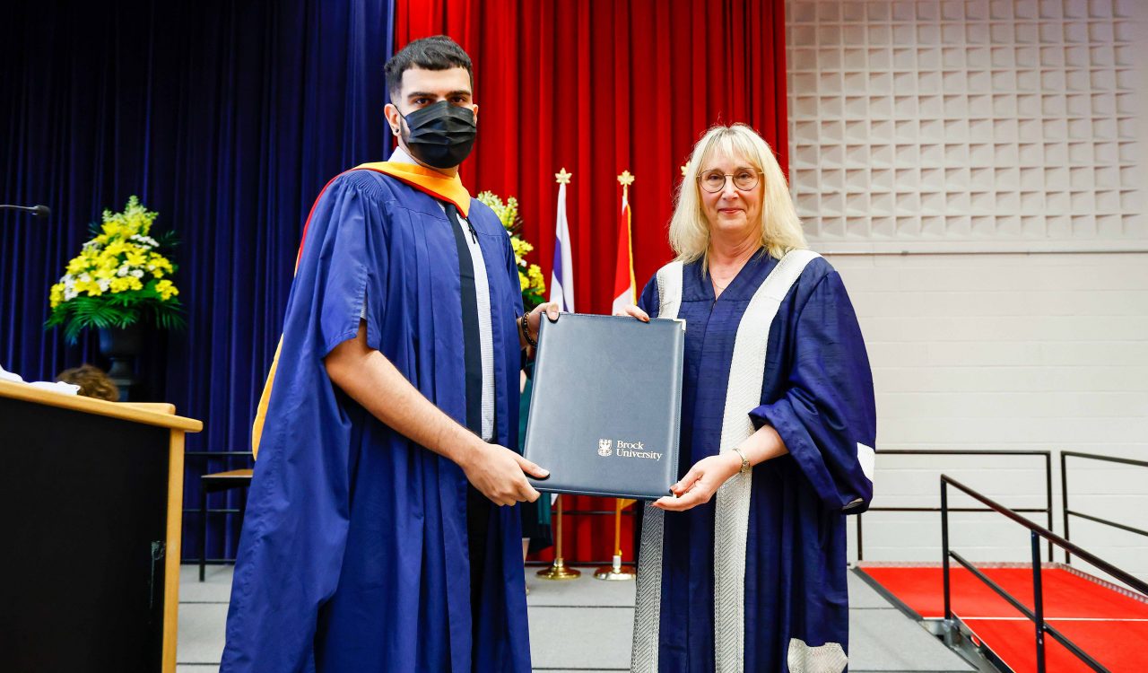 A man and a woman wearing Convocation gowns hold a folder between them as they pose for a photo.