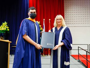 A man and a woman wearing Convocation gowns hold a folder between them as they pose for a photo.