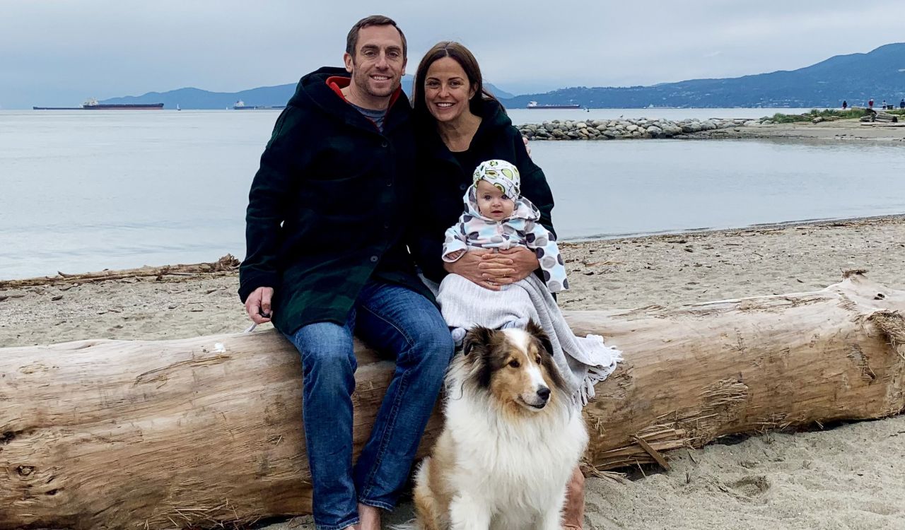 A father and mother sit on a large log in front of the ocean with their baby on mom's lap and their dog sitting at their feet.