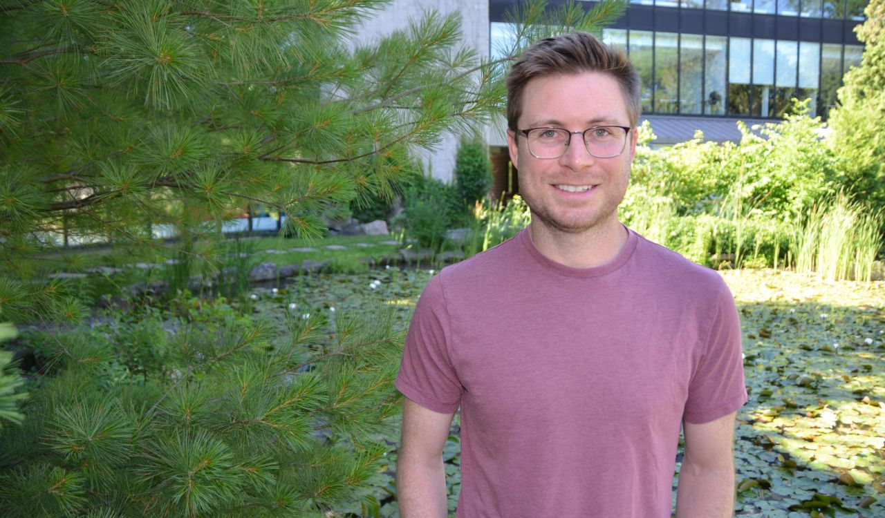 A smiling man with short brown hair and glasses wearing a pinkish T-shirt stands in front of trees with a pond and a building with large glass windows in the background.