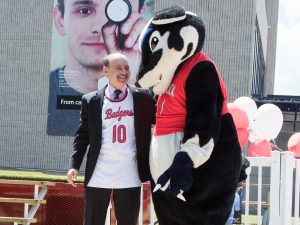 Jack Lightstone, wearing a white and red Brock Badgers No. 10 basketball jersey under a black suit jacket, smiles as he wraps one arm around a large badger mascot wearing a red basketball jersey.