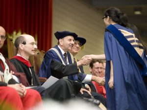 Jack Lightstone shakes the hand of a Brock University student as she walks across the Convocation stage. Seated next to Lightstone are several others, all dressed in ceremonial regalia.