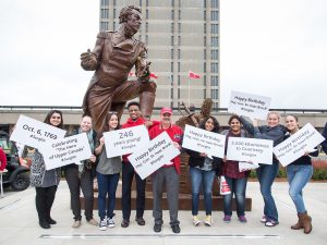 Nine people stand in front of a statue of Maj.-Gen. Sir Isaac Brock. They hold celebratory signs in honour of his 246th birthday.