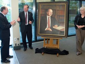Three people stand next to a large oil painting of Jack Lightstone. A black sheet has fallen to the floor below the painting.