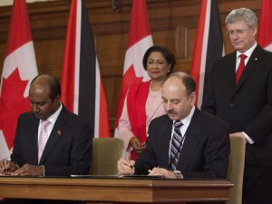 Two men sit at a table signing paper documents with a pen. A woman and a man stand behind them. In the background, Canadian and Trinidadian flags stand against the walls.