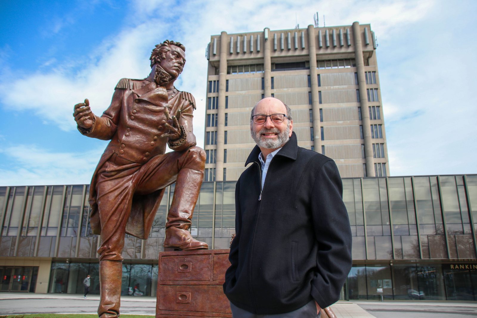 A man stands in front of a large bronze statue of Maj.-Gen. Sir Isaac Brock and Brock University's Schmon Tower.