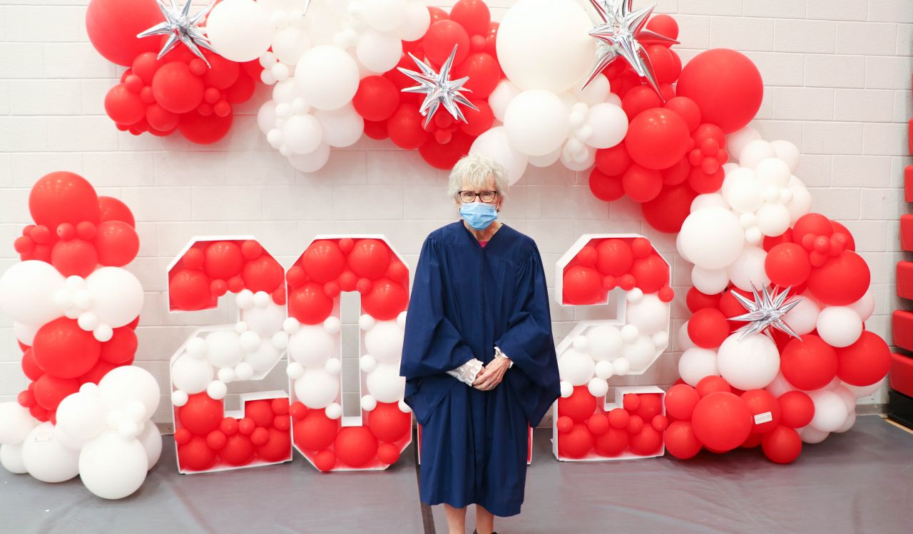An older woman with short white hair stands in a blue graduation gown with white and red balloons in the background, some of which spell out '2022.'