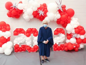 An older woman with short white hair stands in a blue graduation gown with white and red balloons in the background, some of which spell out '2022.'