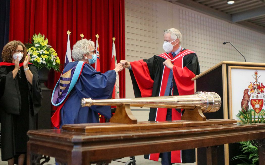A woman with white hair fist bumps a man on stage. Both are wearing graduation gowns.