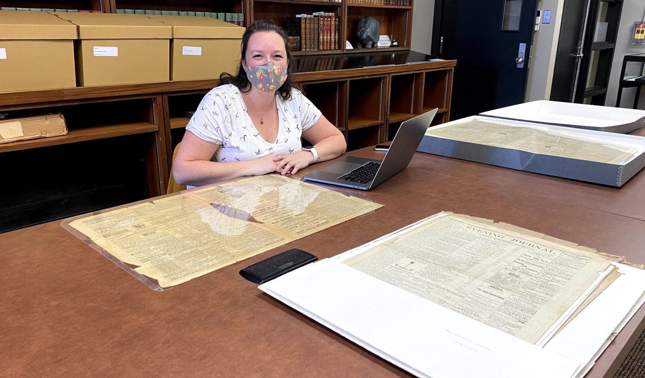 Jessie Richards sits at a table with an archival print artifact in front of her, along with a laptop and several other artifacts beside her. She is sitting in front of bookshelves stacked with rare books and collection pieces in the background.