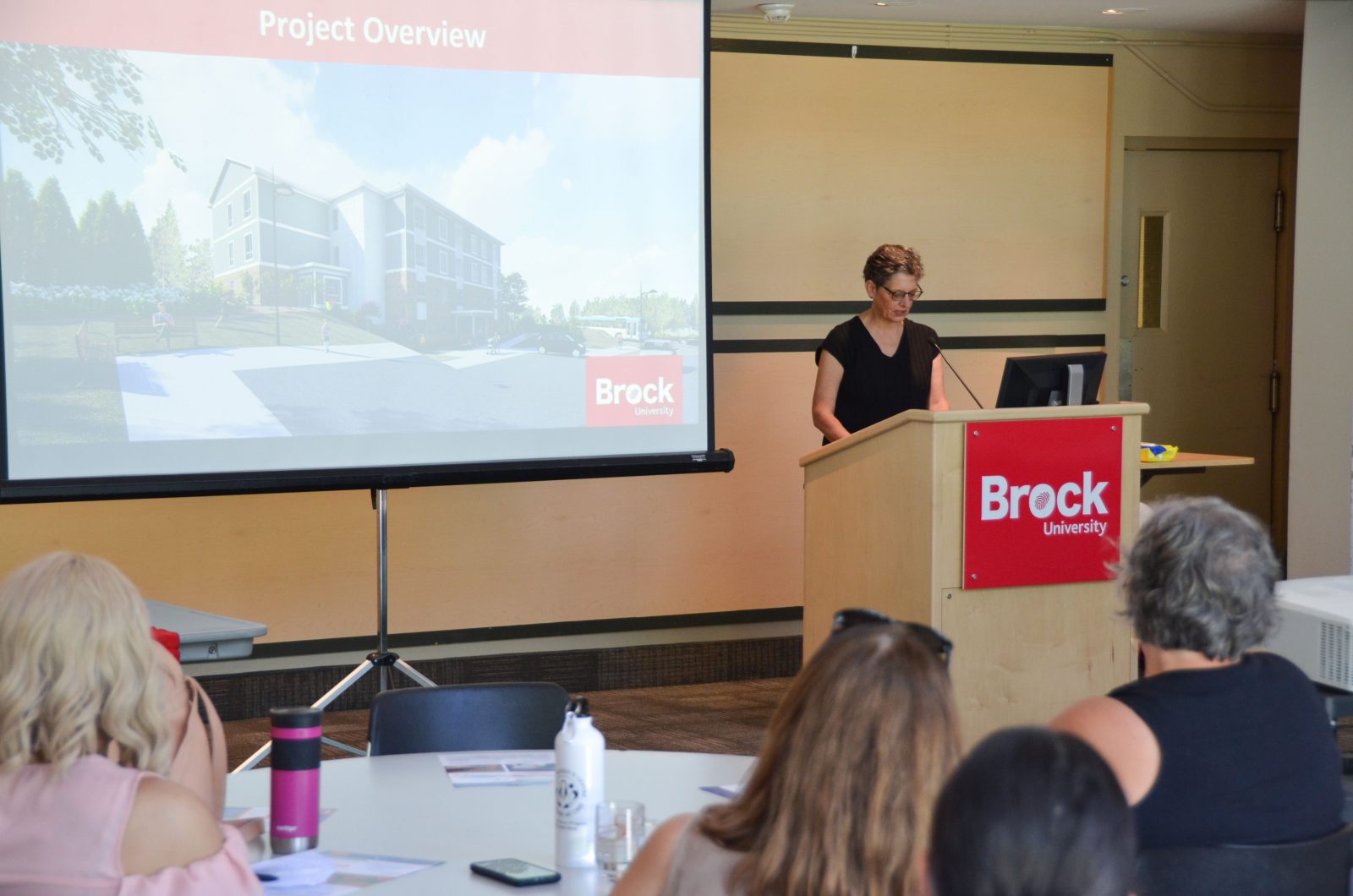 A woman with glasses and wearing a black blouse stands behind a wooden podium labelled Brock University speaking into a microphone. To the left is a slide displayed on a screen. People's heads listening appear in the foreground.