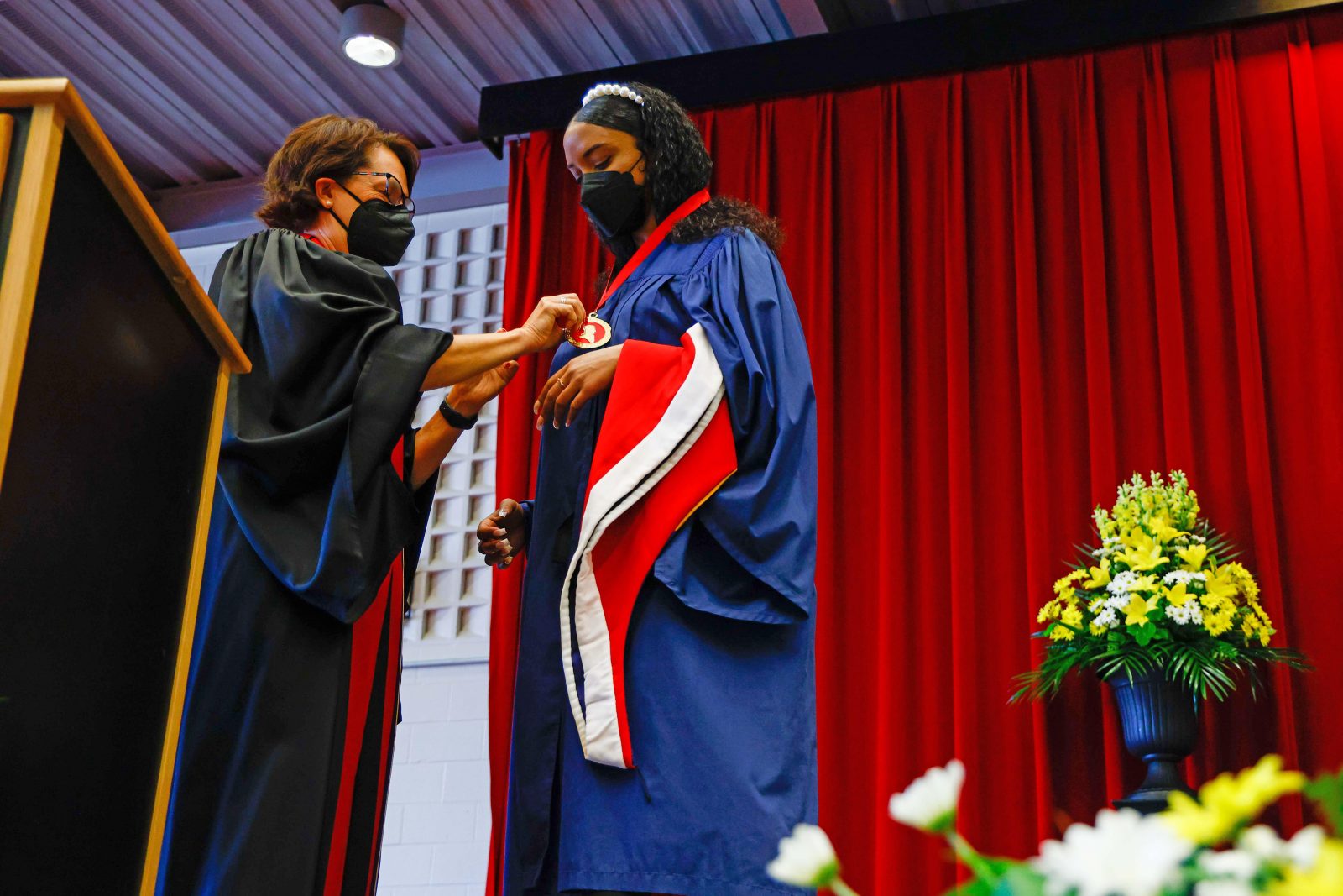 A woman puts a medal around another woman's neck. Both stand in front of a red curtain.