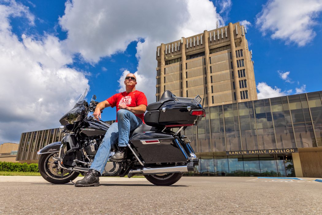 A man sits on a motorcycle in front of a tall concrete tower and wide glass-walled building.