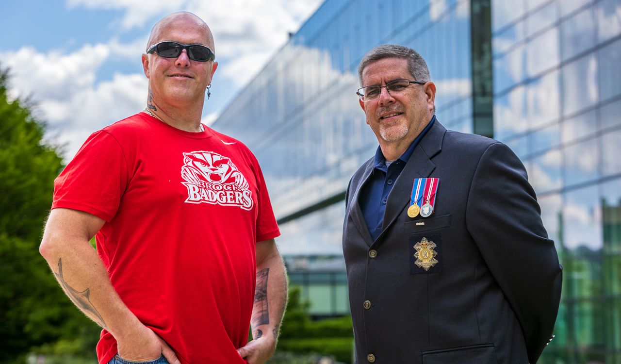 Two men stand outside next to each other in front of a large glass building. The man to the left is wearing a red Brock Badgers T-shirt and sunglasses. The man to the right is wearing a navy suit jacket displaying military medals.