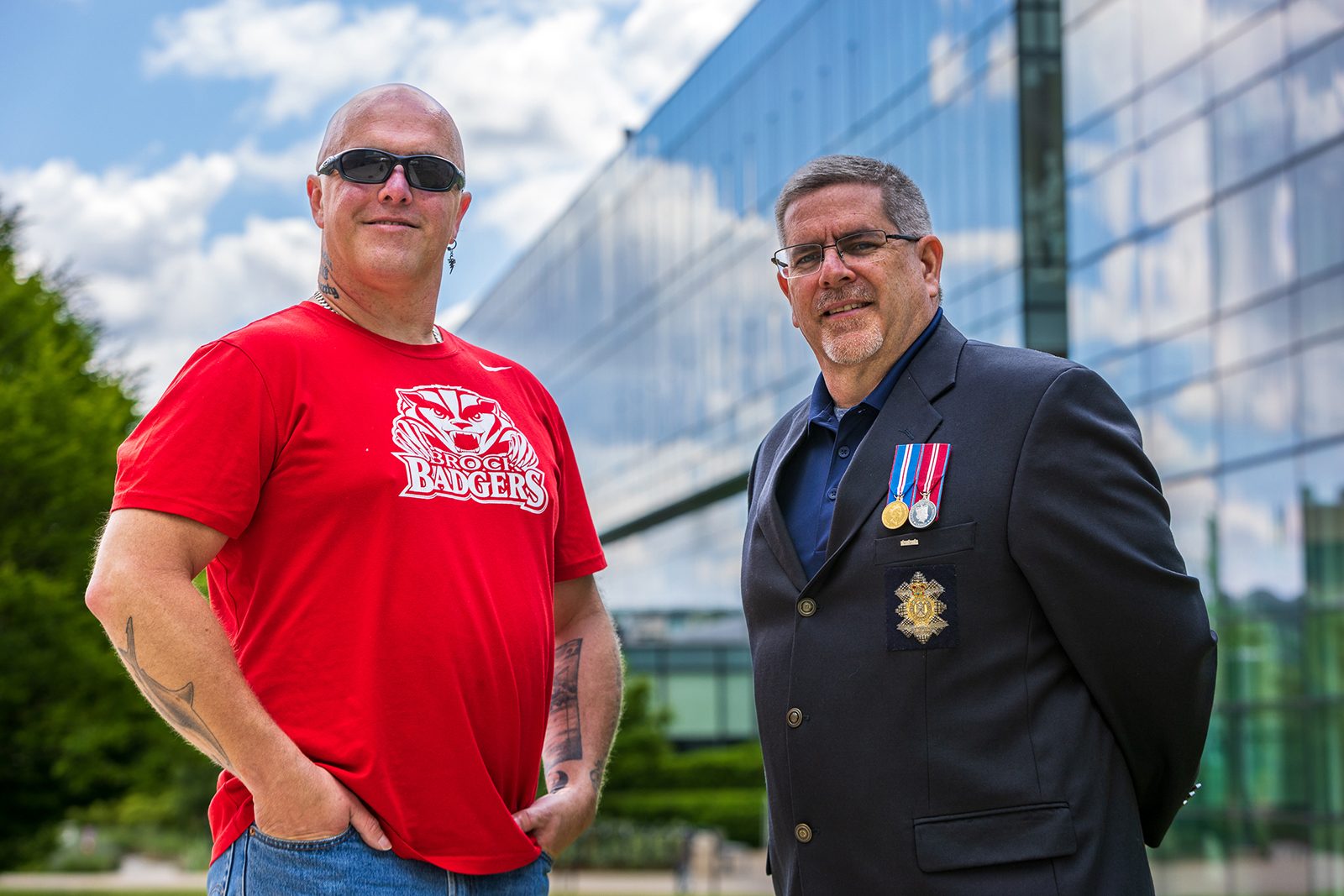 Two men stand outside next to each other in front of a large glass building. The man to the left is wearing a red Brock Badgers T-shirt and sunglasses. The man to the right is wearing a navy suit jacket displaying military medals.