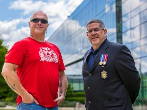 Two men stand outside next to each other in front of a large glass building. The man to the left is wearing a red Brock Badgers T-shirt and sunglasses. The man to the right is wearing a navy suit jacket displaying military medals.