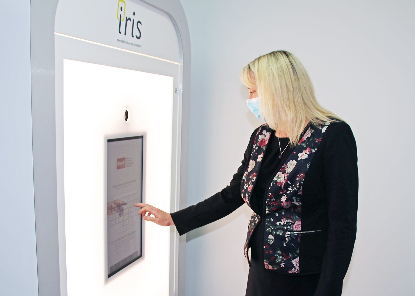 A woman stands in front of a large digital screen that is part of the Iris professional photo booth. She reaches her right hand to the screen to enter contact information.