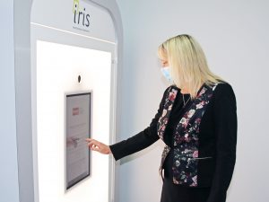 A woman stands in front of a large digital screen that is part of the Iris professional photo booth. She reaches her right hand to the screen to enter contact information.