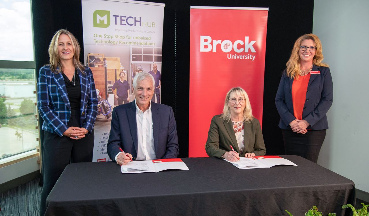 Two people are seated at a table, each with a person standing to their side. Banners for MTechHub and Brock University are featured in the background.