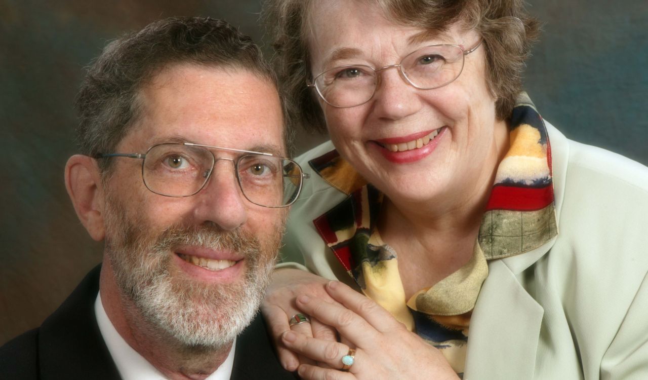 A man and a woman pose together in front of a beige backdrop.