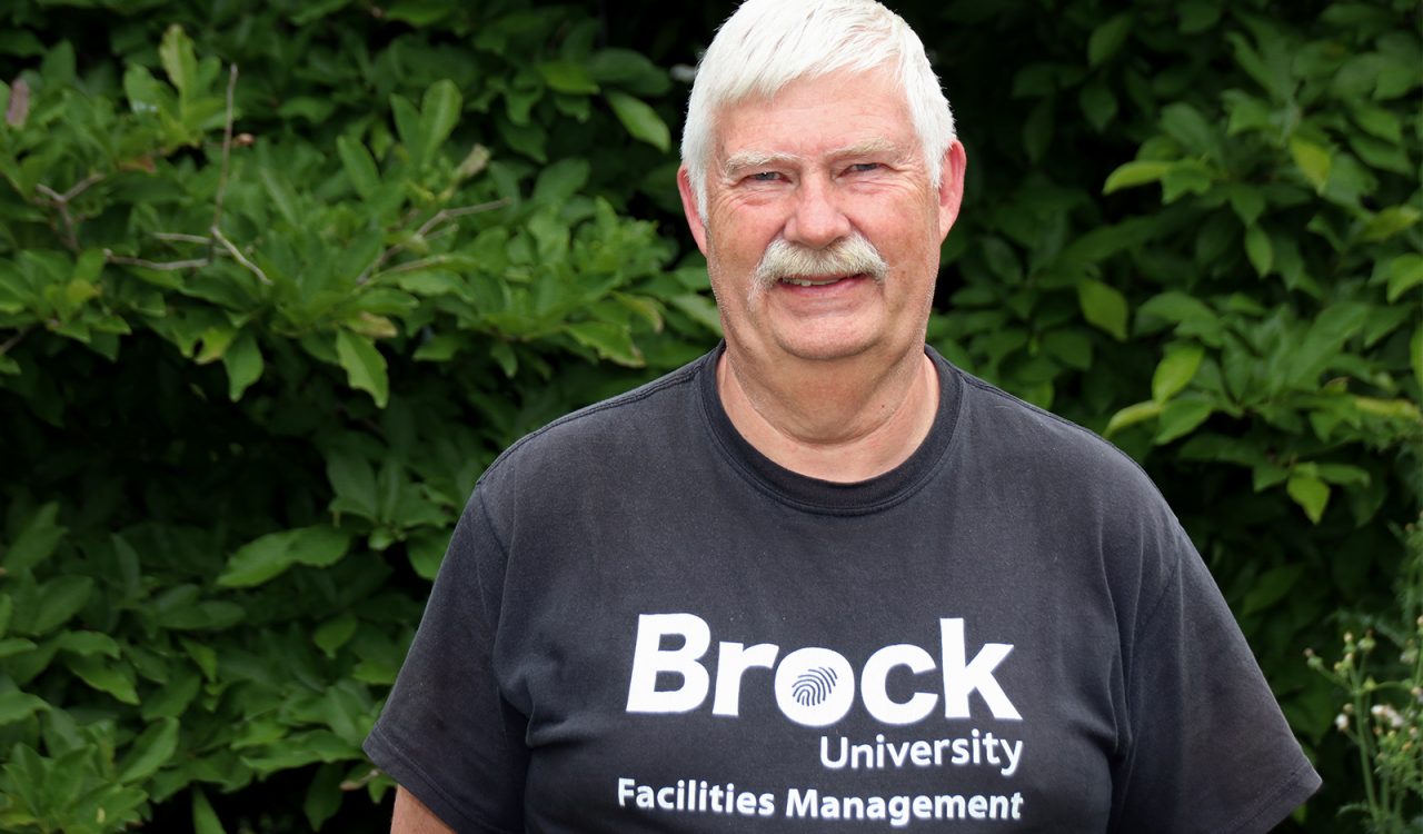 A man wearing a black T-shirt with ‘Brock University Facilities Management’ written on it stands in front of a tall green bush.