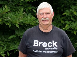 A man wearing a black T-shirt with ‘Brock University Facilities Management’ written on it stands in front of a tall green bush.