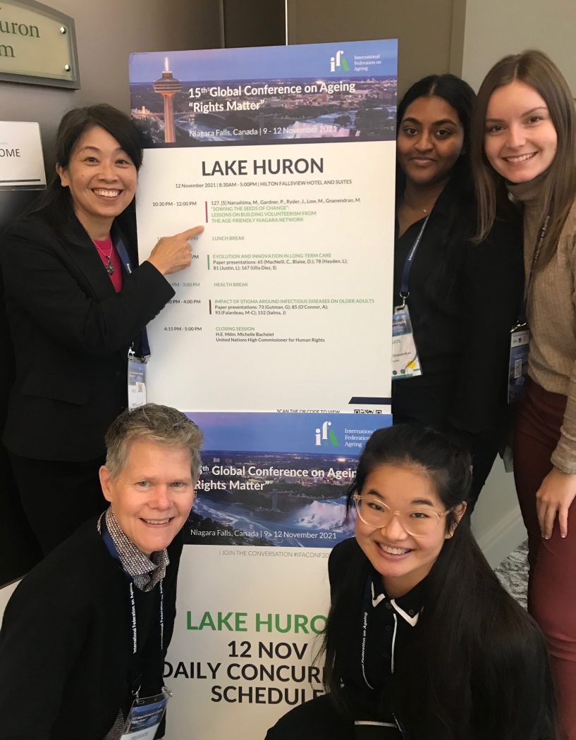 Five women (three standing, two squatting) on both sides of a pop-up banner titled “Lake Huron,” with the woman on the top left pointing at the banner.
