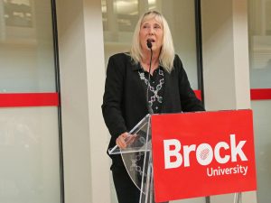 A person speaks into a microphone at a podium with a sign that reads “Brock University”