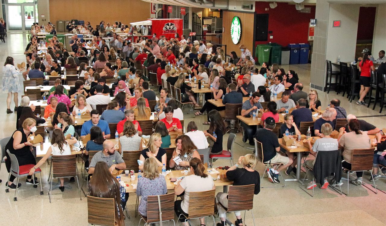 A large, naturally lit indoor space with many tables and chairs filled with people eating lunch.