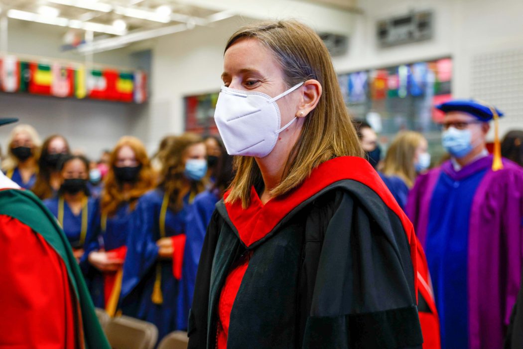 A woman in a mask and graduation gown stands in front of a crowd of people.