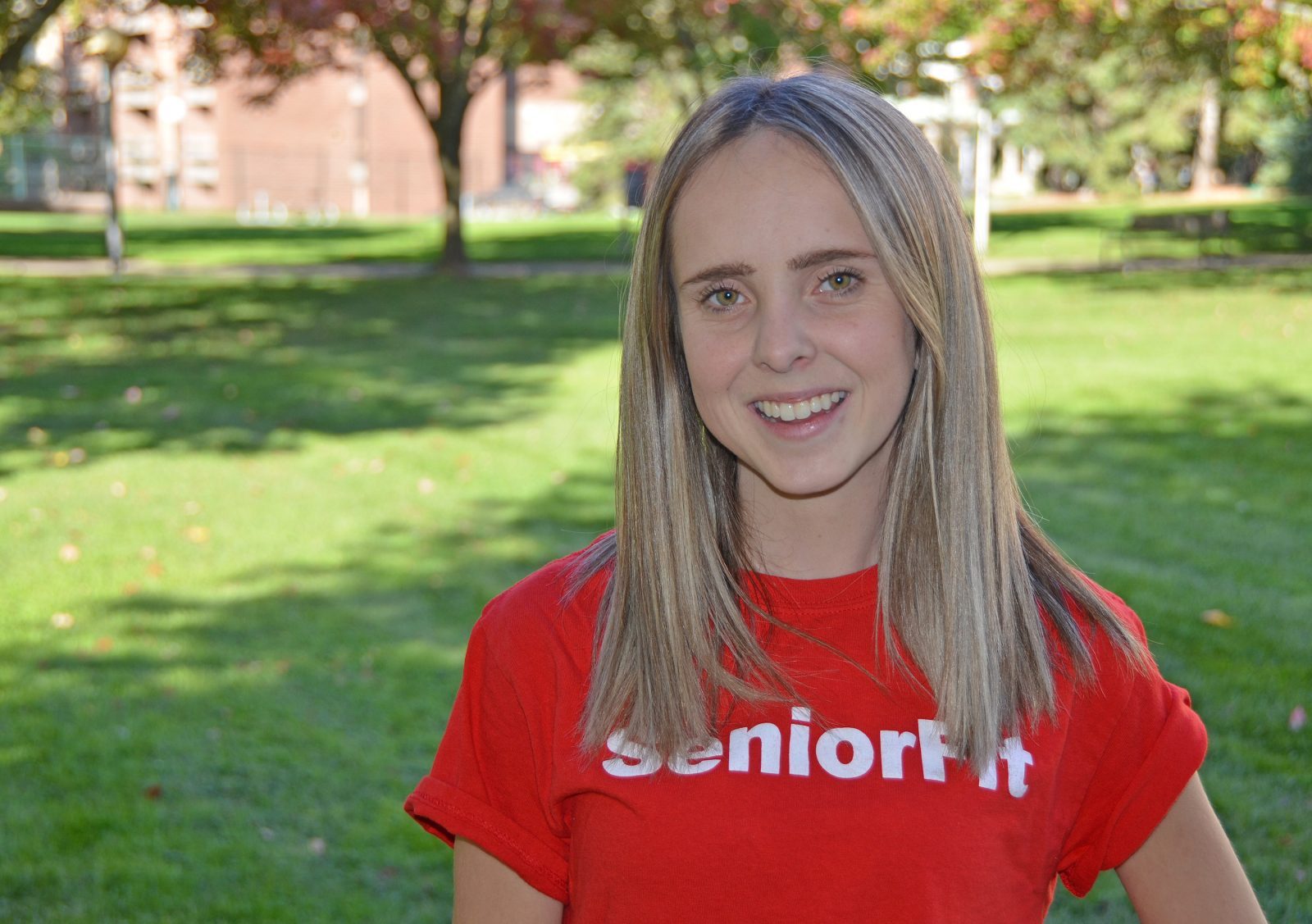 A young, smiling woman with long blond hair wearing a red T-shirt that says “SeniorFit” smiles into the camera, with blurred trees and a building in the background.