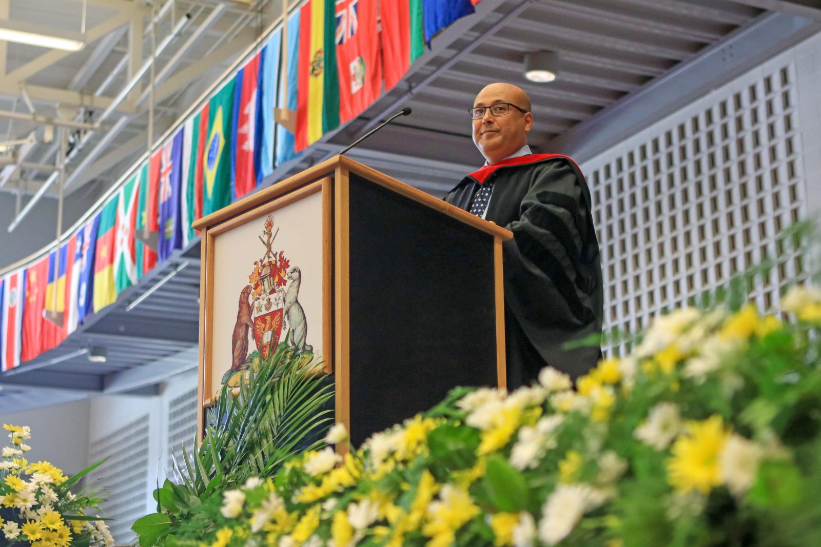 A man stands at a podium in a Convocation gown with yellow flowers in the foreground.