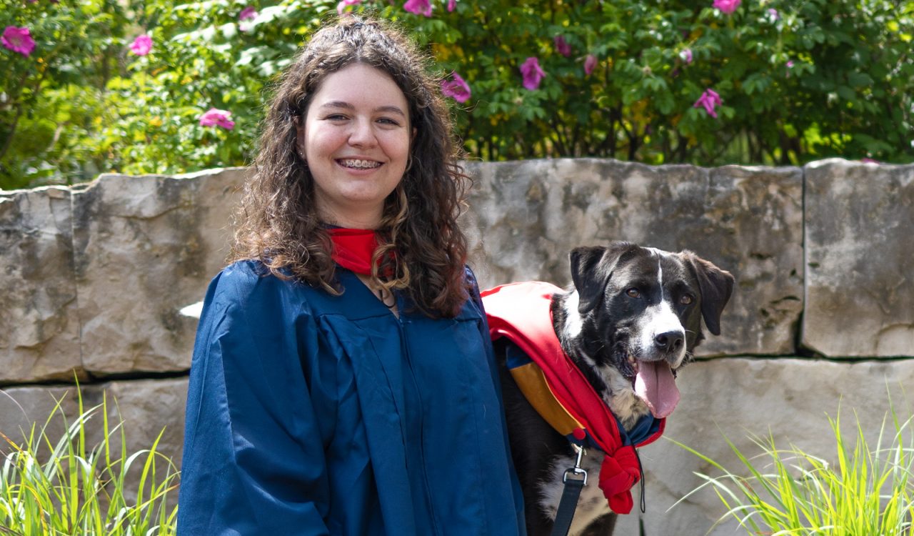 A woman in a graduation gown poses beside a dog, also in a gown.