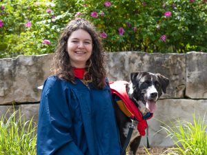 A woman in a graduation gown poses beside a dog, also in a gown.