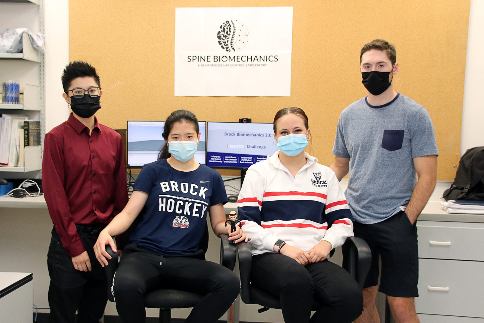 Four young adults stand or sit next to each other in front of a computer screen that reads ‘Brock Biomechanics 2.0, Xsens Challenge’ and a poster that reads ‘Spine Biomechanics and Neuromuscular Control Laboratory.’