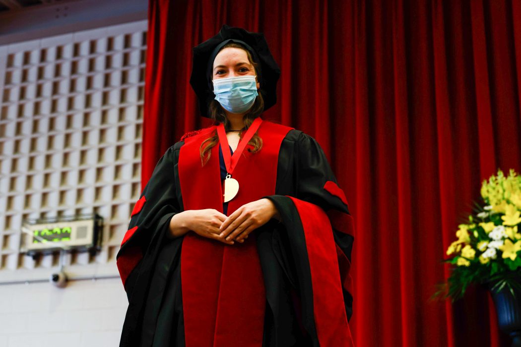 A woman in a graduation gown and hat with a medal around her neck stands in front of red curtains.