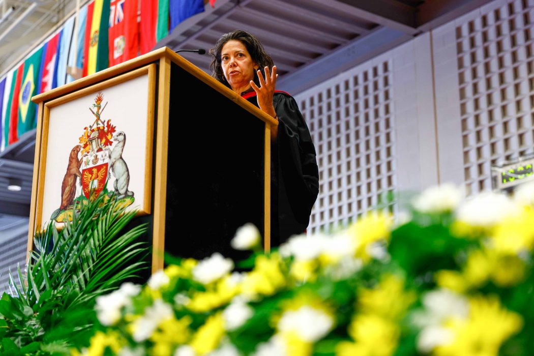 A woman stands behind a podium speaking with yellow and white flowers in the foreground.