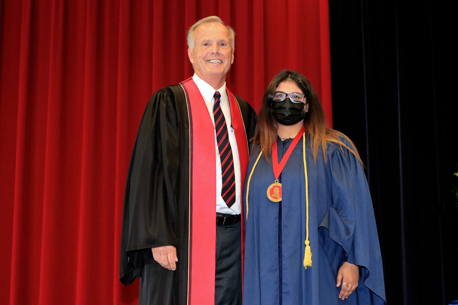 A man and a woman in Convocation gowns stand together with red and black curtains in the background.