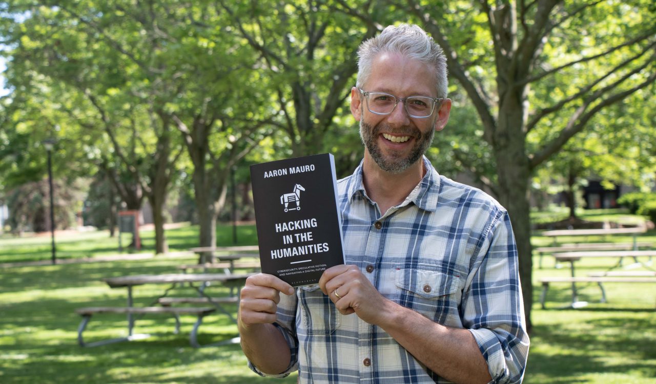 Aaron Mauro, with a white and blue plaid shirt on, holds a copy of his book in his hands while standing outdoors in a treed courtyard.