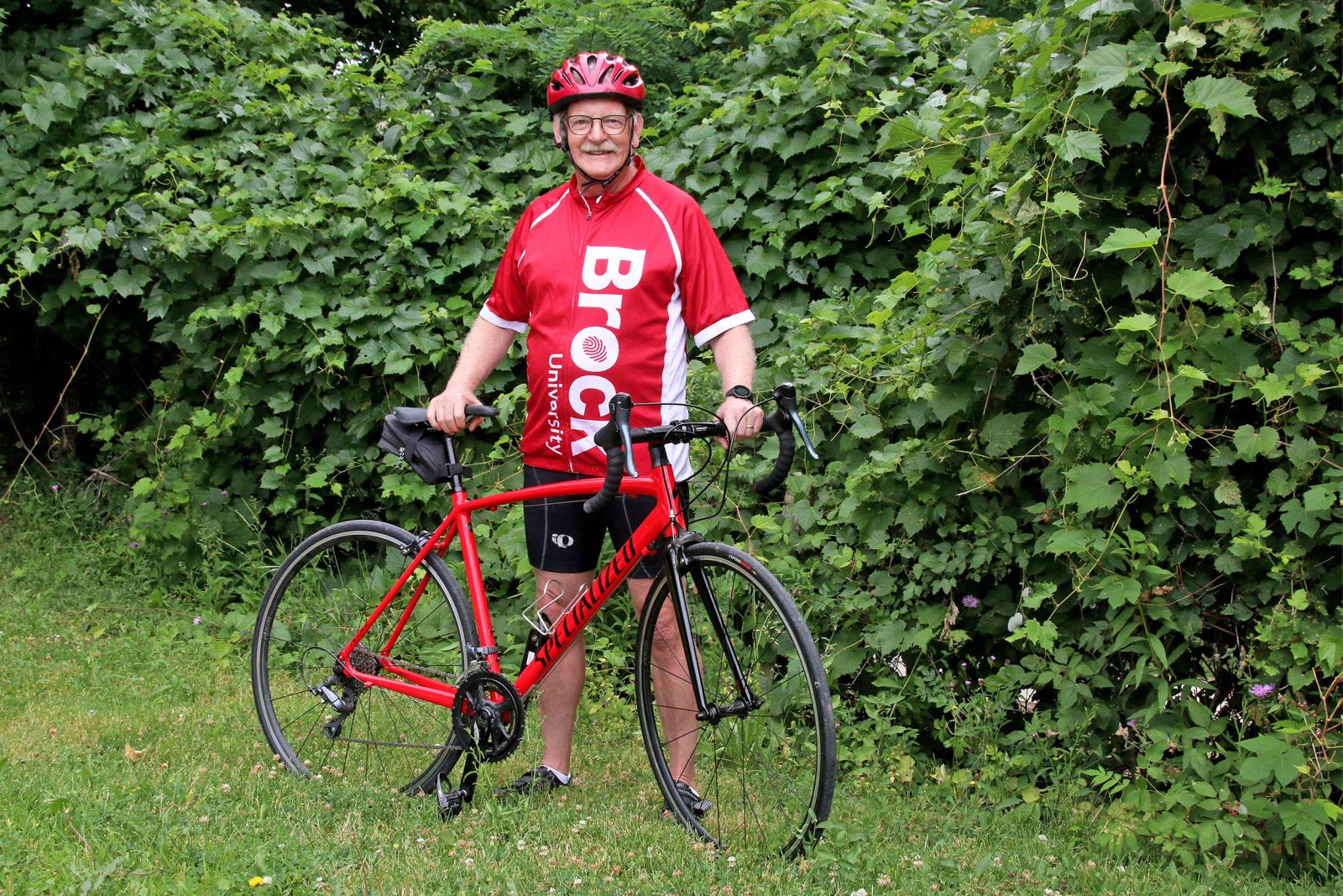 Alan Castle poses in front of lush greenery with his red bicycle. He is wearing a red Brock University cycling jersey, a red helmet and black cycling shoes.