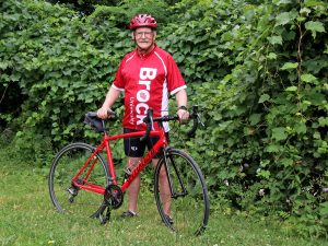 Alan Castle poses in front of lush greenery with his red bicycle. He is wearing a red Brock University cycling jersey, a red helmet and black cycling shoes.